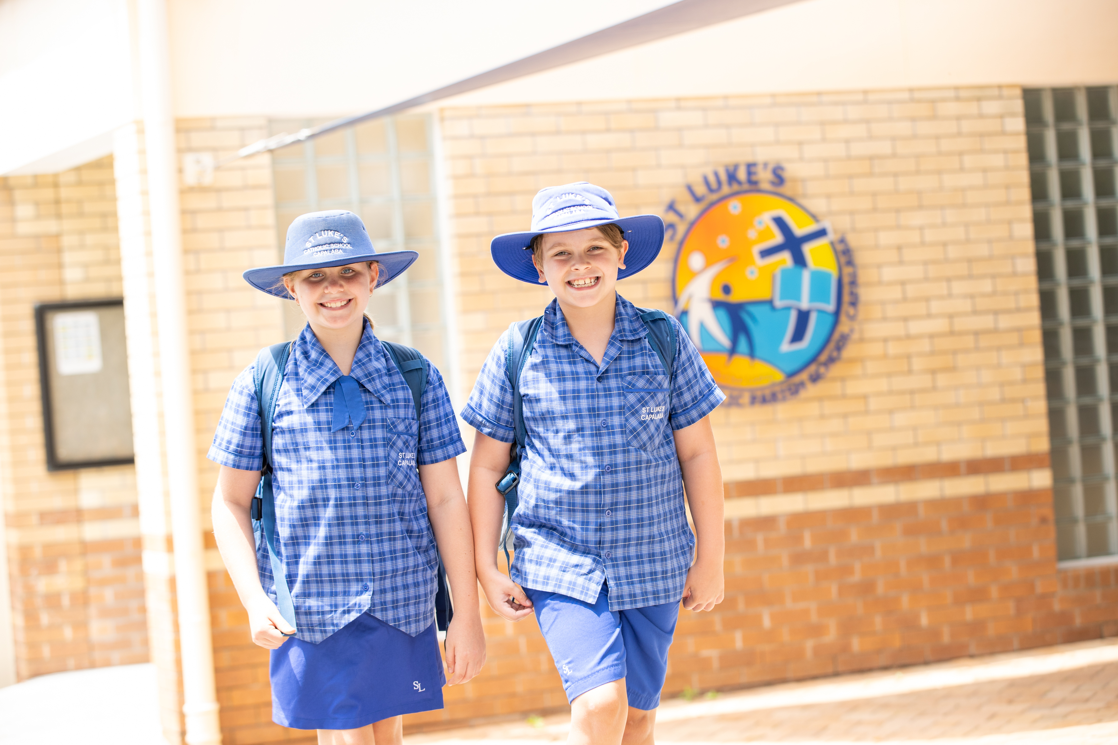 Two students in school uniform walking outdoors past a brick school building with a St Luke’s school emblem on the wall.