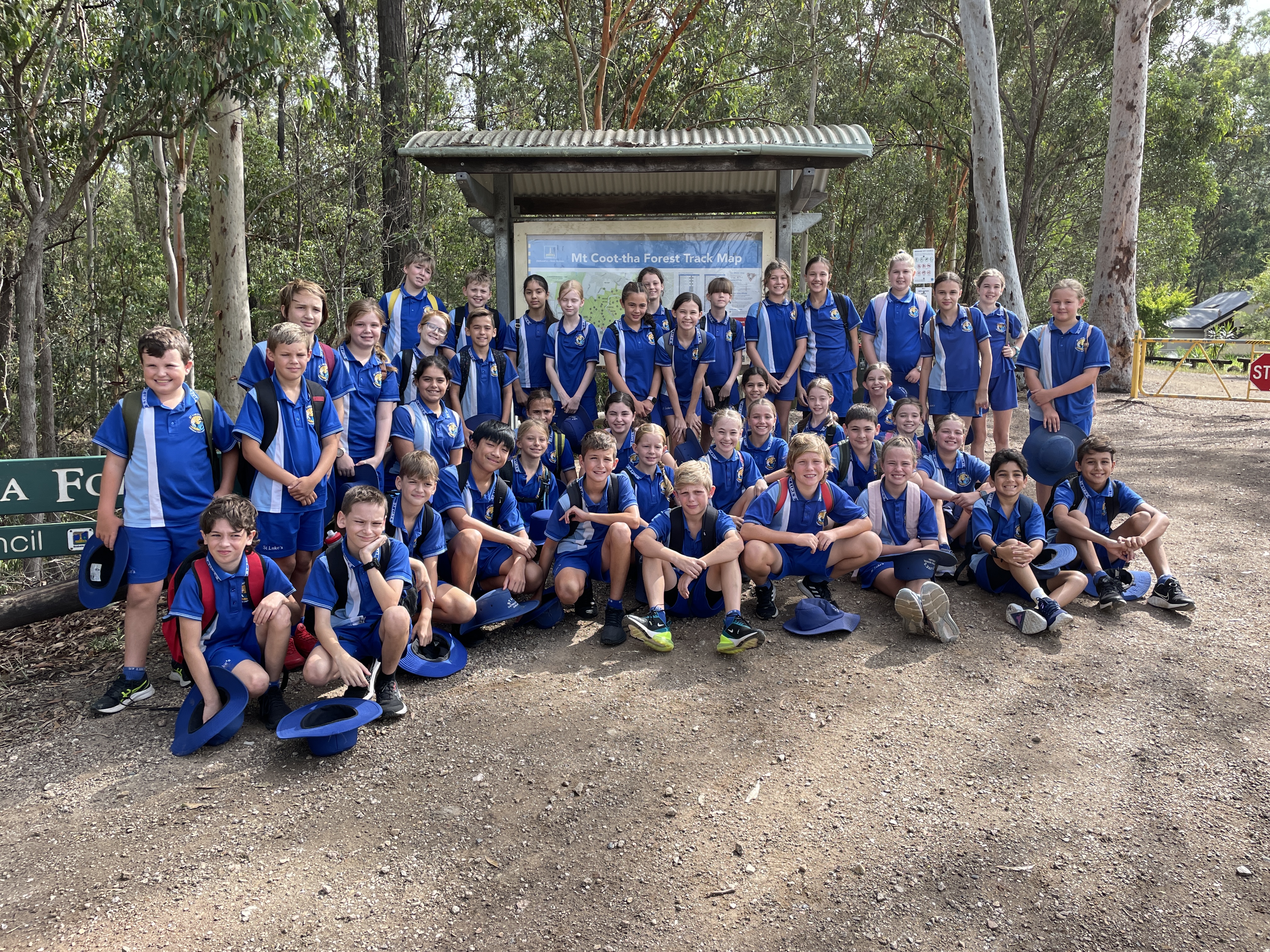 Large group of students in school sports uniforms gathered outdoors in front of a forest track map during a school excursion.