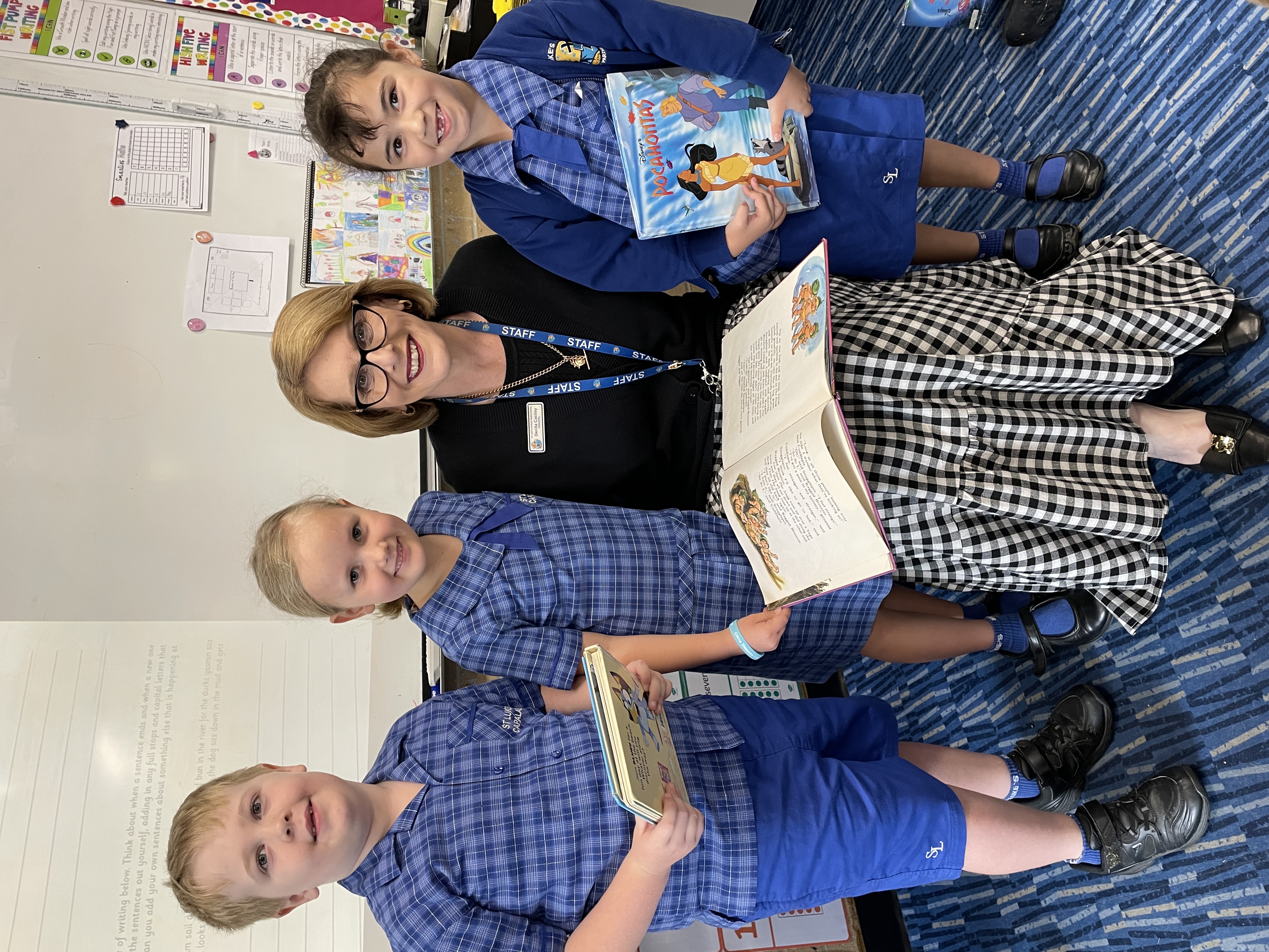 Staff member seated in a classroom holding an open book, with three students in school uniform standing nearby holding books.