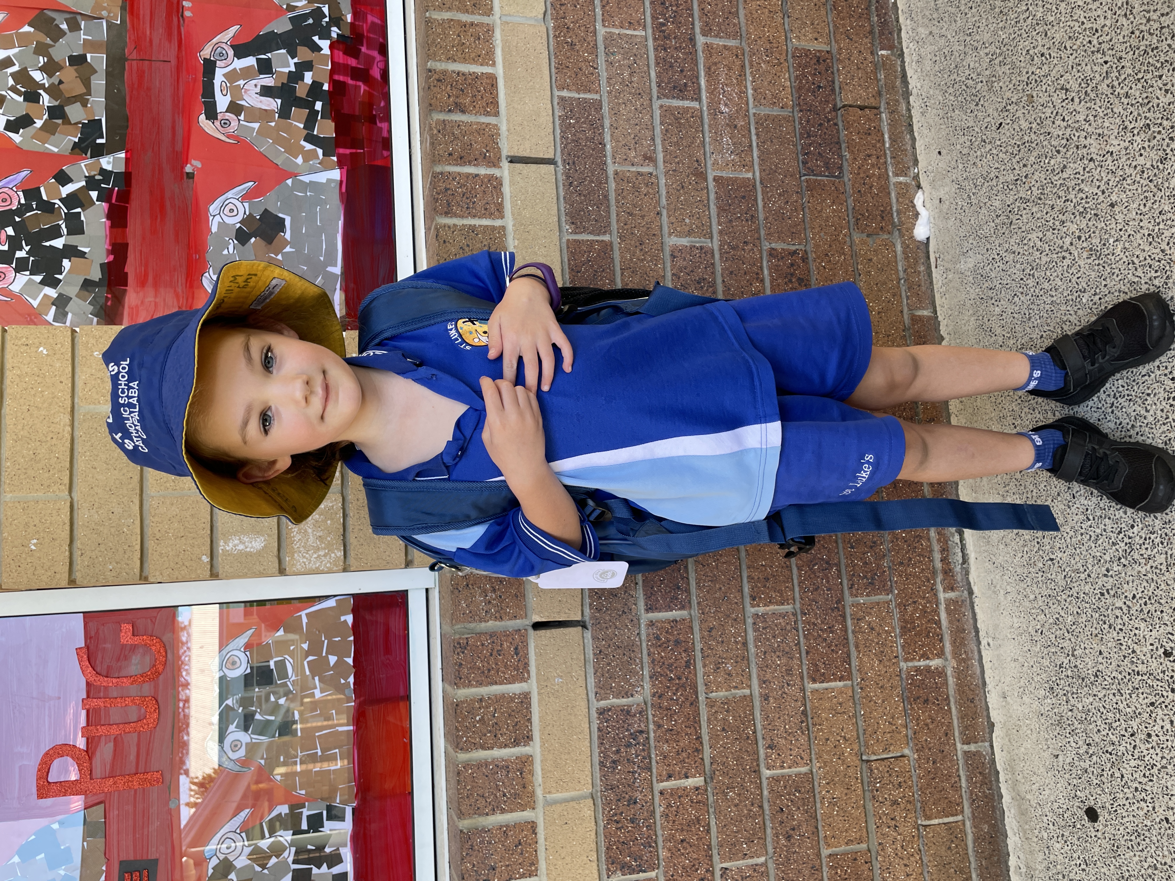 Student in school uniform standing outdoors with a backpack, in front of a classroom window.