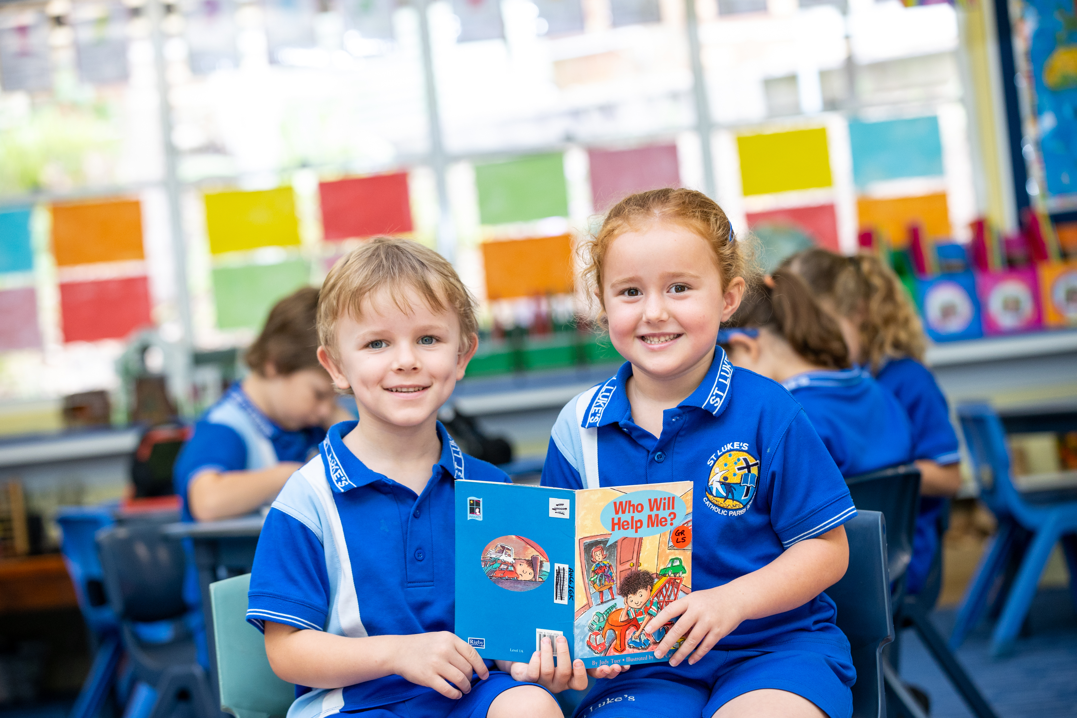 Two students in school uniform sitting together in a classroom holding an open children’s book.