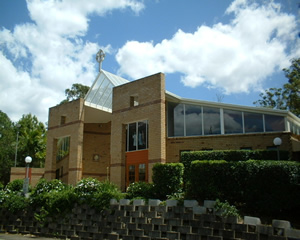 Brick church building with a cross on the roof and large windows, surrounded by greenery under a blue sky.