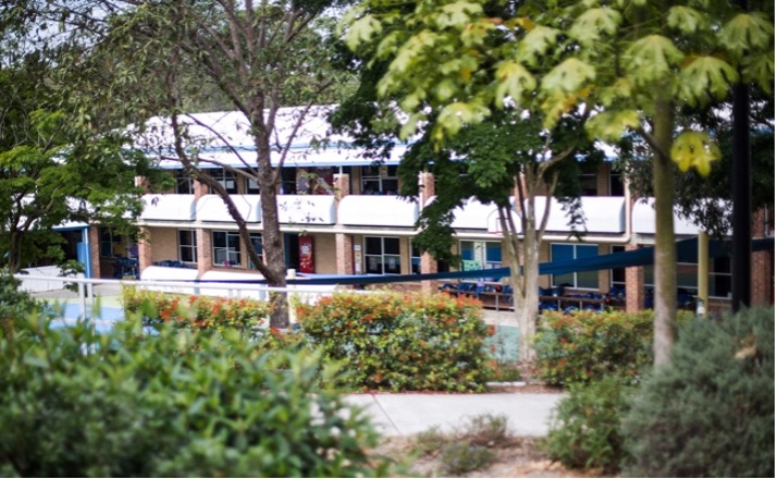 School campus building with classrooms and walkways, viewed across landscaped gardens and trees.